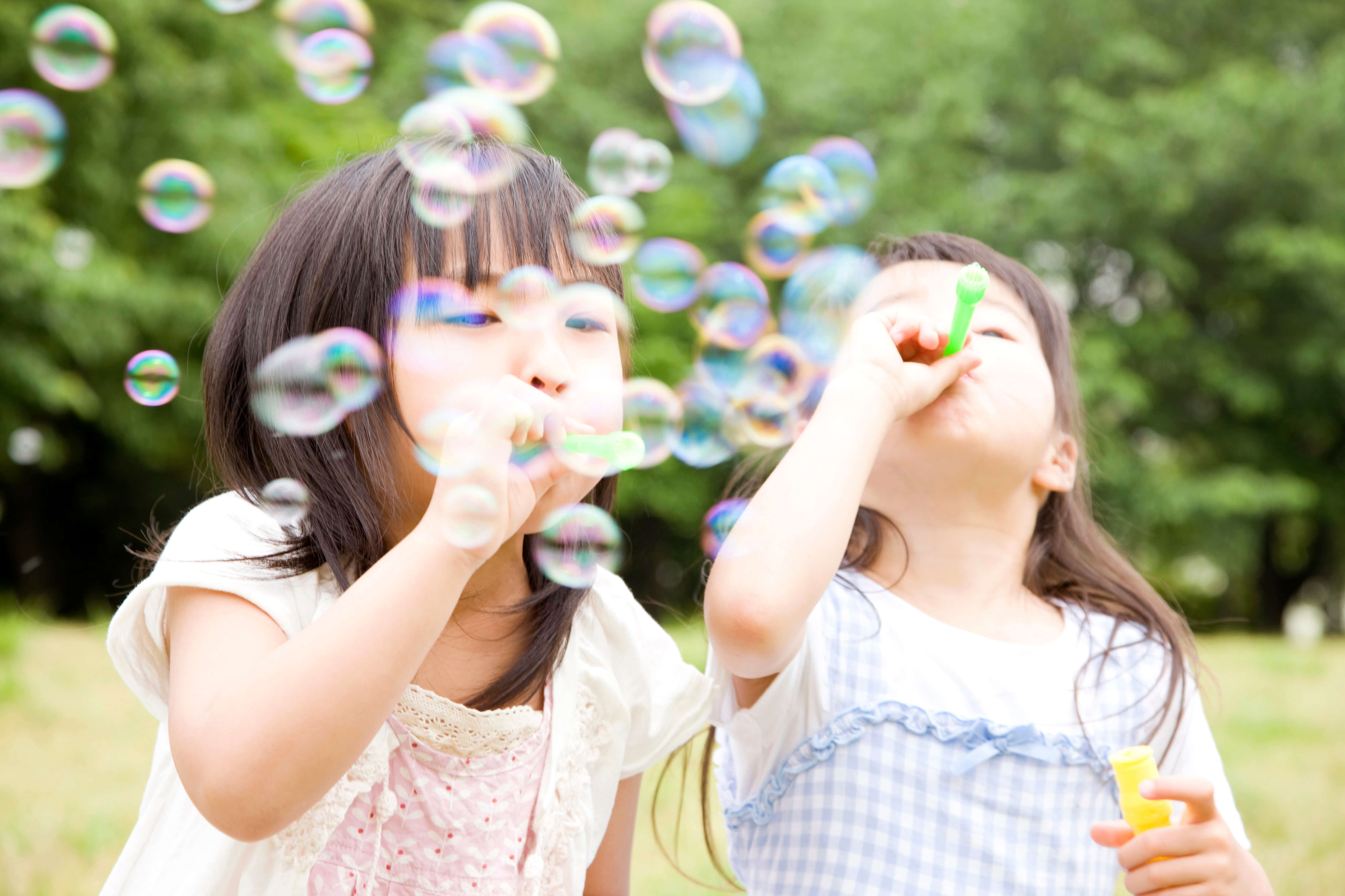 緑豊かな屋外で、シャボン玉を吹いて遊ぶ白い服を着た二人の子供。たくさんのシャボン玉が空中を舞う瞬間