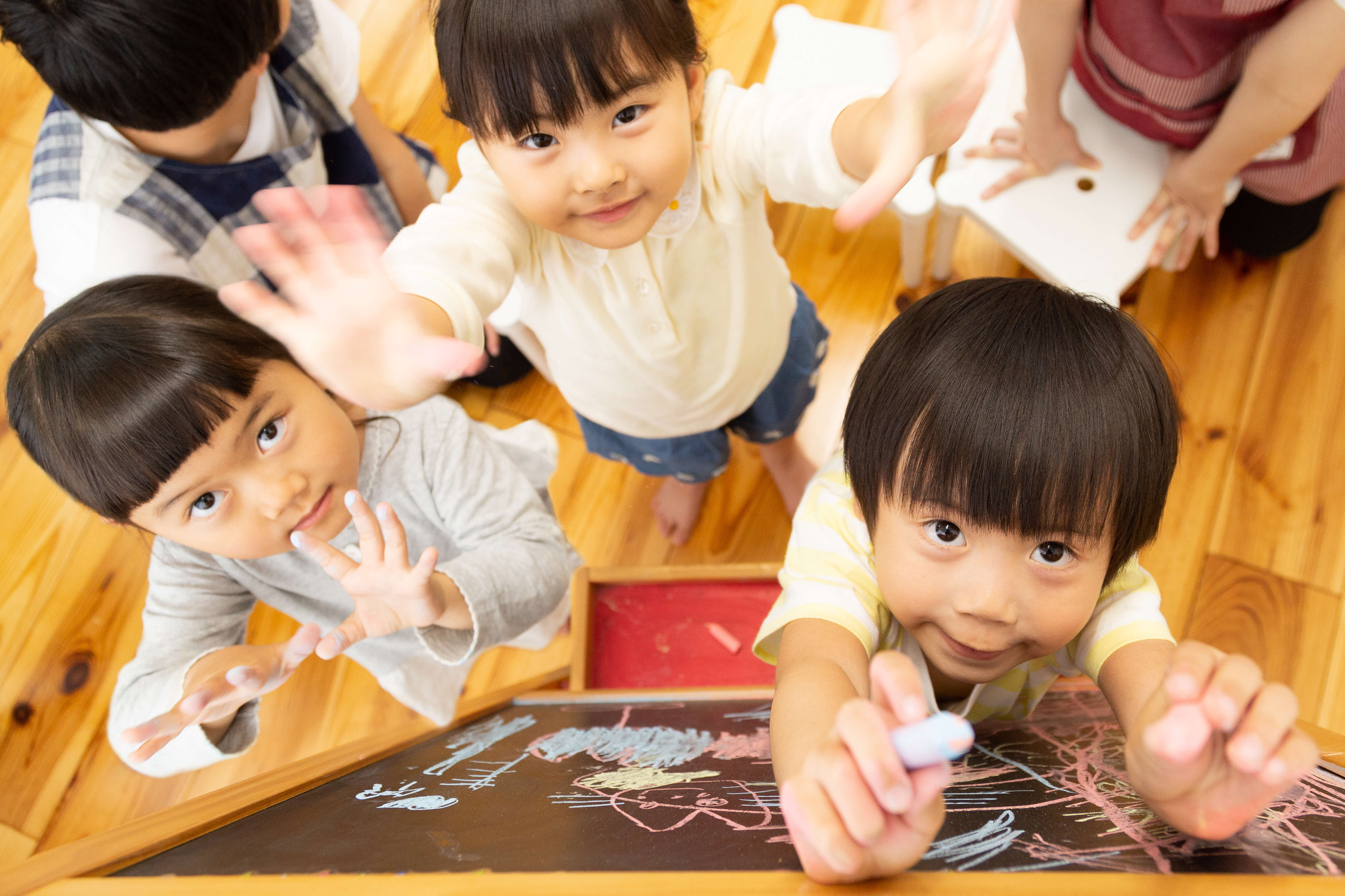 木製の床で描画や工作を楽しむ子供たち。上から撮影された明るい笑顔の様子